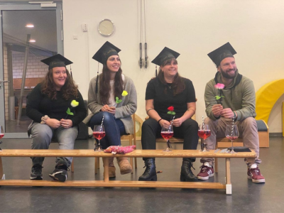 Vier Personen sitzen nebeneinander auf kleinen Stühlen in einem Gruppenraum. Alle tragen schwarze Doktorhüte und halten je eine farbige Blume in der Hand. Vor ihnen stehen Gläser mit rotem Getränk auf einer Holzbank. Die Atmosphäre wirkt festlich und konzentriert.