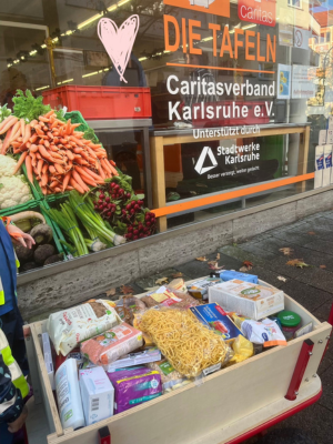 Holzwagen mit gespendeten Lebensmitteln wie Nudeln, Reis und Konserven vor dem Schaufenster der Karlsruher Tafel. Hinter dem Glas sind Gemüseauslagen und das Logo des Caritasverbands Karlsruhe e. V. zu sehen.