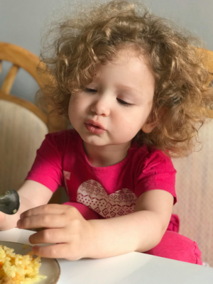 Kleines Kind sitzt am Tisch und greift selbstständig nach Essen auf dem Teller, Blick nach unten gerichtet.