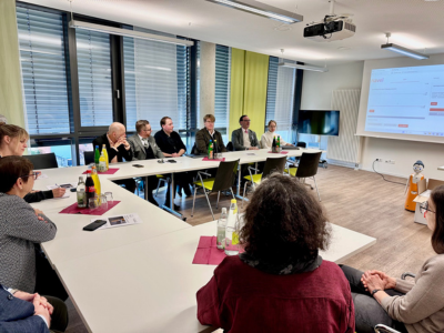Personengruppe sitzt in einem Besprechungsraum an einem großen, U-förmigen Tisch. Vorne im Bild mehrere Glasflaschen und Notizmaterial, im Hintergrund Präsentationsfolie auf einem Bildschirm. Rechts am Rand steht der kleine Roboter „Navel/Felix“.