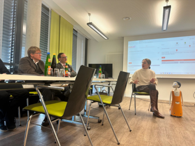 Sophia Warneke sitzt in einem Besprechungsraum auf einem Stuhl und spricht. Neben ihr steht der orangefarbene Roboter „Navel/Felix“. Zwei Personen am Tisch hören zu, eine Präsentation ist an die Wand projiziert.