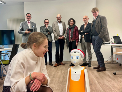 Sophia Warneke kniet vorne im Bild und lächelt den Roboter „Navel/Felix“ an, der eine rote Mütze trägt. Hinter ihr stehen sechs Personen nebeneinander und blicken in Richtung Kamera.