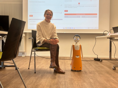 Sophia Warneke sitzt seitlich neben dem Roboter „Navel/Felix“ auf einem Stuhl im Besprechungsraum. Hinter beiden läuft eine Präsentation über einen Beamer an der Wand.