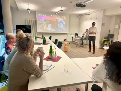 Mehrere Personen sitzen in einem Besprechungsraum an einem U-Tisch. Vorderseite Kaffee und Getränke, im Hintergrund Sophia Warneke stehend vor einer Präsentationsfolie. Rechts steht der Roboter „Navel/Felix“.