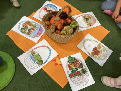 Korb mit Obst und Gemüse in der Mitte eines orangenen Tuchs. Rundherum liegen von Kindern gestaltete Bilder mit ausgemalten Erntekörben. Kinderbeine sind am Rand sichtbar.