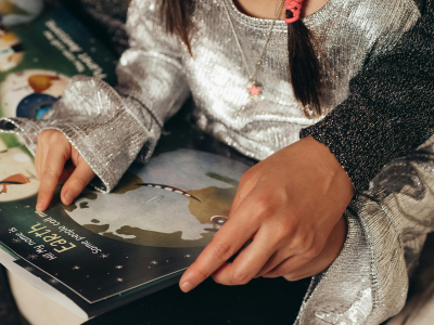Ein Kind und eine erwachsene Person blättern gemeinsam in einem farbigen Bilderbuch. Beide tragen glitzernde Kleidung, die Hände liegen nebeneinander auf der aufgeschlagenen Seite.