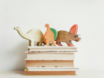 Drei bunte Holz-Dinosaurier stehen auf einem Stapel Kinderbücher vor einer hellen Wand. Die Figuren sind dekorativ nebeneinander angeordnet.
