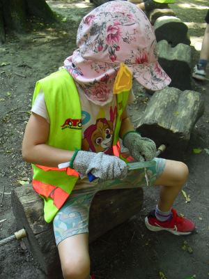 Ein Kind mit Warnweste, Handschuhen und Sonnenhut sitzt auf einem Holzbalken und schnitzt konzentriert an einem Stück Holz.