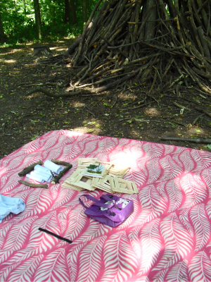 Picknickdecke im Wald mit Holzbausteinen, Körben und Materialien für Kreativangebote. Rechts steht eine große Hütte aus Ästen.
