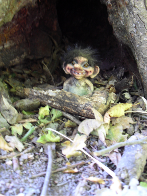 Ein kleiner Trollfigur-Kopf schaut aus einem hohlen Baumstamm heraus, umgeben von Laub und Holzstücken.