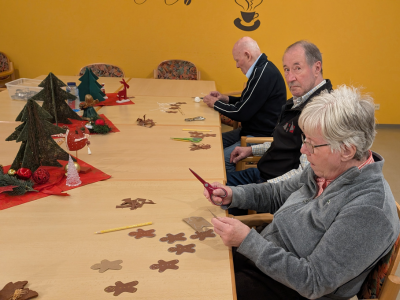 Mehrere Bewohner*innen sitzen an einem langen Tisch und schneiden aus braunem Teig Sterne und andere Formen. Auf dem Tisch stehen kleine Weihnachtsdekorationen.