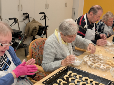Bewohner*innen tragen Schürzen und formen gemeinsam Vanillekipferl. Auf dem Tisch stehen Schüsseln, Puderzucker und ein Backblech mit Gebäck.