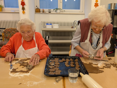 Zwei Bewohnerinnen sitzen nebeneinander und stechen mit Förmchen Plätzchen aus Teig aus. Vor ihnen liegt ein Backblech mit vorbereiteten Plätzchen.