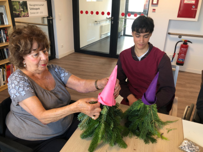 Eine Seniorin und ein junger Mann basteln gemeinsam an zwei Weihnachtswichteln aus Tannenzweigen. Beide setzen pinke und lilafarbene Filzmützen auf die kleinen Figuren.