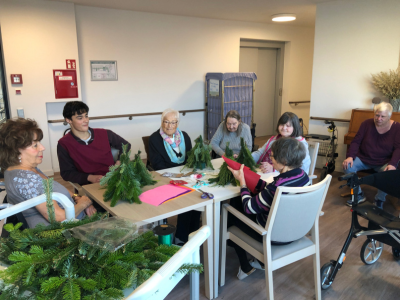 Eine Gruppe von sieben Personen sitzt an einem Tisch und bastelt Weihnachtswichtel aus frischen Tannenzweigen. Auf dem Tisch liegen Bastelmaterialien wie Filz, Draht und rote Pompoms.