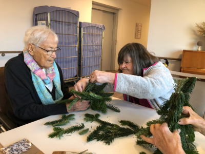 Zwei Seniorinnen arbeiten konzentriert gemeinsam an einem Tannenwichtel. Eine hält den Zweig fest, während die andere ihn sorgfältig befestigt.