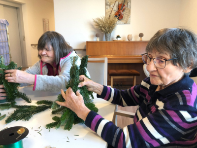 Zwei Frauen befestigen Tannenzweige zu kleinen Weihnachtsfiguren. Im Hintergrund steht ein Klavier, im Vordergrund liegen Draht und Bastelmaterial.
