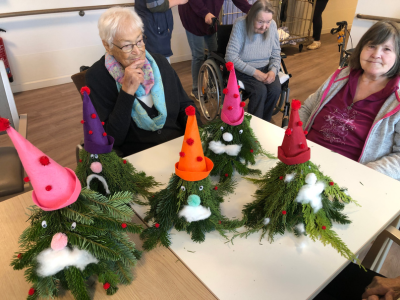 Sechs fertige Wichtel mit bunten Zipfelmützen, Bärten aus Watte und roten Nasen stehen auf einem Tisch. Zwei Bewohnerinnen sitzen stolz daneben.