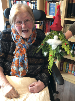 Eine Seniorin im Rollstuhl hält lächelnd einen fertigen Tannenwichtel mit roter Mütze und weißem Bart auf dem Arm. Im Hintergrund sieht man ein Bücherregal.