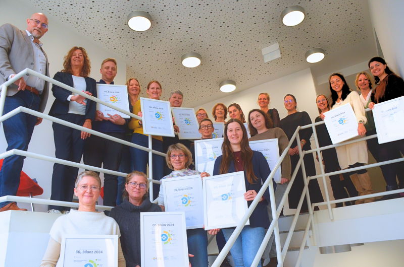 Gruppenfoto der Leitungen der AWO-Kindertagesstätten auf einer Treppe in der Geschäftsstelle. Sie halten ihre CO₂-Zertifikate in den Händen. Links stehen Markus Barton, Geschäftsführer der AWO Karlsruhe, und Dorothée Eby aus dem Klimaschutzmanagement. In der oberen Mitte sind Esther Marggrander, Geschäftsbereichsleitung Kitas, und Lucie Adamek aus dem Klimaschutzmanagement zu sehen.
