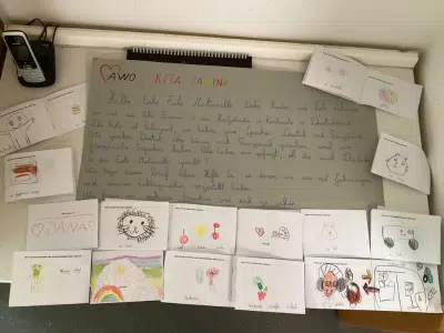 Ein großes graues Plakat mit handgeschriebenem Text und mehreren Kinderzeichnungen liegt auf einem Tisch. Rundherum sind gefaltete Karten mit bunten Zeichnungen der Kinder ausgelegt. Links oben liegt ein Telefon.