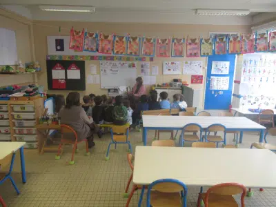 Eine Gruppe von Kindern sitzt in einem Klassenraum auf kleinen Stühlen im Halbkreis vor einer Tafel. Eine erwachsene Person sitzt daneben. Die Wände sind mit Kinderkunstwerken, Fotos und Lernmaterialien dekoriert.