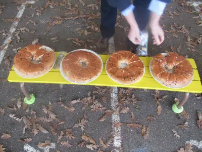 Vier große runde Hefekränze liegen auf Tellern auf einer gelben Bank im Freien. Eine Person richtet die Teller aus. Auf dem Boden liegen viele trockene Eichenblätter.