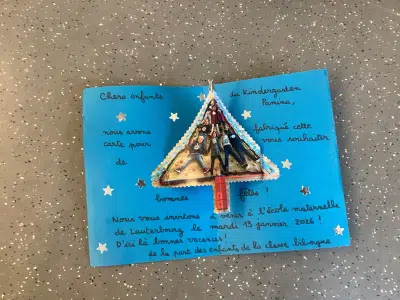 Eine blaue Karte mit französischem Text und aufgeklebt gebasteltem Weihnachtsbaum liegt auf dem Boden. Der Baum besteht aus Papier, Glitzer und kleinen Dekoelementen.
