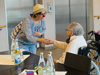 Ein Clown in gestreiftem Shirt, Latzhose und roter Nase begrüßt freundlich eine Seniorin im Rollstuhl. Die beiden halten sich an den Händen und lächeln einander an. Auf dem Tisch stehen mehrere Wasserflaschen und Gläser. Die Begegnung vermittelt Nähe und Freude.