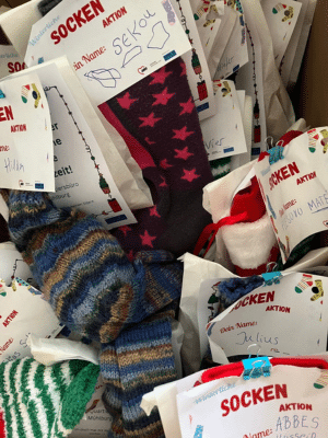 Ein Korb ist gefüllt mit bunten, handgestrickten Socken. An vielen Paaren hängen handbeschriftete Etiketten mit dem Wort „Socken“ und kleinen Zeichnungen. Die Socken unterscheiden sich in Farben und Mustern. Das Bild zeigt eine vorbereitete Spenden- oder Geschenkaktion.