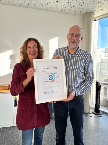 Zwei lächelnde Personen stehen in einem hellen Büro und halten gemeinsam eine gerahmte Urkunde in die Kamera. Auf dem Zertifikat steht groß der Titel „CO2 Bilanz 2024“, ergänzt durch ein kreisförmiges Infografik-Logo und verschiedene Daten zur Emissionsberechnung. Im Hintergrund ist ein modernes Büro mit Fensterfront und ein Aktenregal zu erkennen.