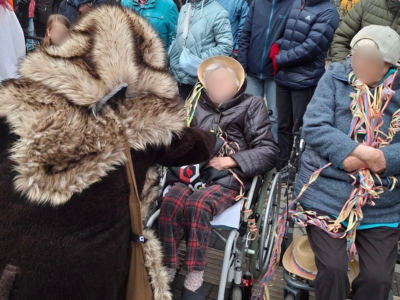 Bewohner*innen des Hauses am Blumentor sitzen beim Faschingsumzug in Rollstühlen. Eine als Fellfigur verkleidete Person begrüßt eine Bewohnerin. Hinter ihnen steht eine dicht gedrängte Menschenmenge.
