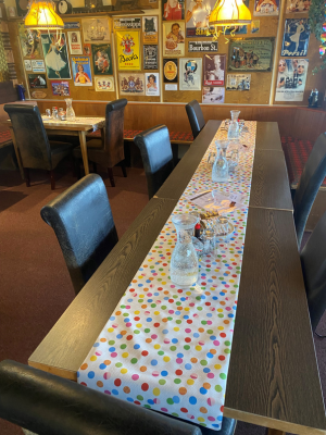 Langer Tisch in einem Restaurant, dekoriert mit einem Faschingsläufer in bunten Punkten. Auf dem Tisch stehen Wasserkaraffen und kleine Schalen mit Snacks. An den Wänden hängen zahlreiche Plakate.