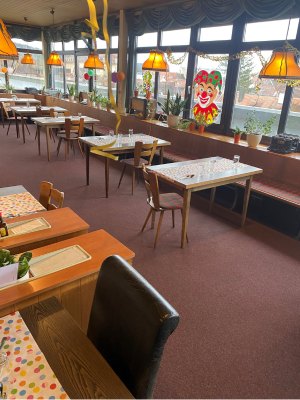 Geschmückter Gastraum mit mehreren Tischen, farbigen Tischläufern und Faschingsdekoration. Durch große Fenster fällt Tageslicht, im Hintergrund hängt ein Clownmotiv an der Scheibe.