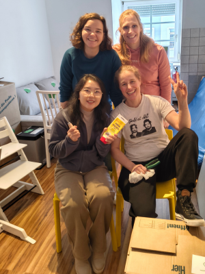 Vier Personen sitzen und stehen dicht beieinander in einem kleinen Raum, lächeln in die Kamera und halten Werkzeug und Material in den Händen.