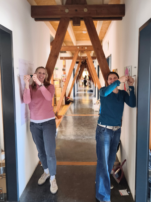 Zwei Personen stehen sich in einem Flur gegenüber und befestigen Papier an den Türrahmen, im Hintergrund ist der lange Gang mit Holzstreben sichtbar.