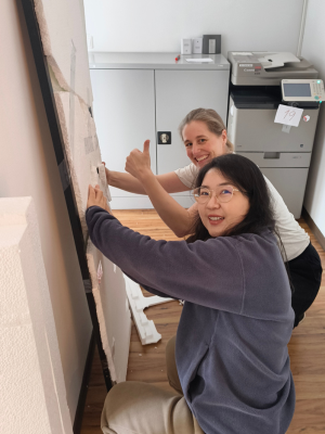 Zwei Personen knien vor einer Wand und arbeiten gemeinsam an einer montierten Fläche, eine Person zeigt einen Daumen hoch, im Hintergrund steht ein Drucker.