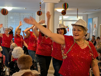 Eine Gruppe von Frauen in roten, glitzernden Kostümen und silbernen Hüten tanzt ausgelassen während der Faschingsfeier.