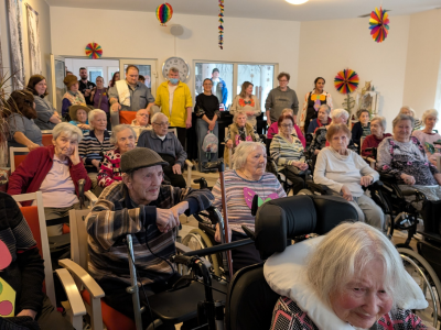 Senior*innen in Rollstühlen und Sitzreihen verfolgen ein Faschingsprogramm im Pflegeheim. Im Hintergrund stehen viele Mitarbeitende und Gäste. Die Stimmung wirkt aufmerksam und lebendig.