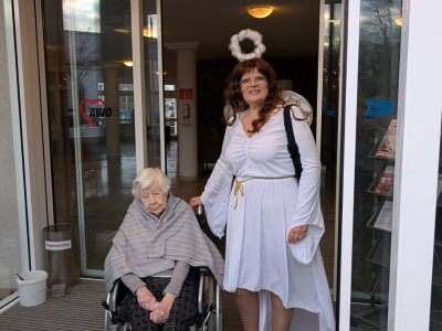 Eine Mitarbeiterin als Engel verkleidet begleitet eine Seniorin im Rollstuhl zum Fest.