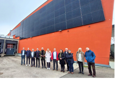 Eine Gruppe von etwa zwölf Personen steht im Außenbereich vor einer großen, orangefarbenen Gebäudewand, an der eine vertikale Photovoltaikanlage montiert ist. Links im Bild sind zwei Laderampen mit Toren zu sehen. Der Himmel ist grau. Die Personen stehen in einer Reihe, einige mit Jacken und Taschen, und blicken in Richtung Kamera.