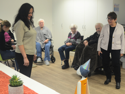 Senior*innen sitzen im Gemeinschaftsraum eines Pflegeheims und beobachten einen orangefarbenen Assistenzroboter mit Partyhut während einer Aktivierungsrunde.