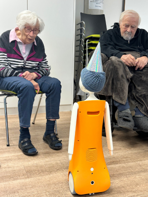 Zwei ältere Bewohner*innen im Pflegeheim sitzen vor einem orangefarbenen sozialen Roboter und reagieren aufmerksam auf dessen Ansprache.