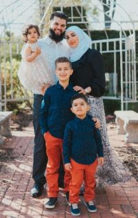 Fünfköpfige Familie steht gemeinsam im Freien vor einer hellen Gartenkulisse. Eltern und drei Kinder posieren nah beieinander für ein Familienfoto.