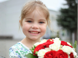 Kleines blondes Mädchen hält einen Strauß roter und weißer Rosen in den Händen und lächelt in die Kamera.
