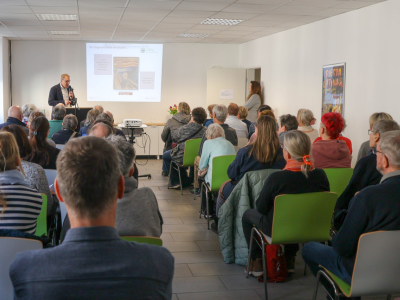 Vortragsraum mit vielen Zuhörer*innen. Eine Präsentation läuft auf der Leinwand, vorne spricht ein Mann.