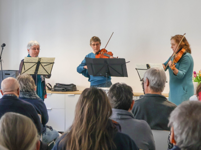 Drei Musikerinnen spielen ein Stück auf Streichinstrumenten und Querflöte vor Publikum.