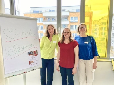 Drei Frauen stehen nebeneinander in einem hellen Raum vor großen Fenstern. Neben ihnen steht ein Flipchart mit der handschriftlichen Aufschrift „Herzlich willkommen“ und einem kleinen Plakat. Die Frauen tragen Namensschilder und lächeln in die Kamera. Im Hintergrund sind Wohngebäude zu sehen, ein Teil des Fensters ist gelb eingefärbt.