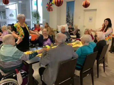 Bewohner*innen und Mitarbeiter*innen in bunten Westen sitzen lachend beisammen und halten sich bei der gemeinsamen Faschingsfeier an den Händen.