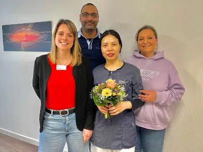 Alt-Text: Vier lächelnde Personen stehen nebeneinander in einem Innenraum vor einer hellen Wand. In der Mitte hält eine Frau in blauer Arbeitskleidung einen bunten Blumenstrauß. Links neben ihr steht eine Frau in rotem Oberteil und schwarzer Strickjacke, rechts eine Frau in einem fliederfarbenen Kapuzenpullover. Hinter der Gruppe steht ein Mann mit Brille. Ein abstraktes Bild hängt links an der Wand.